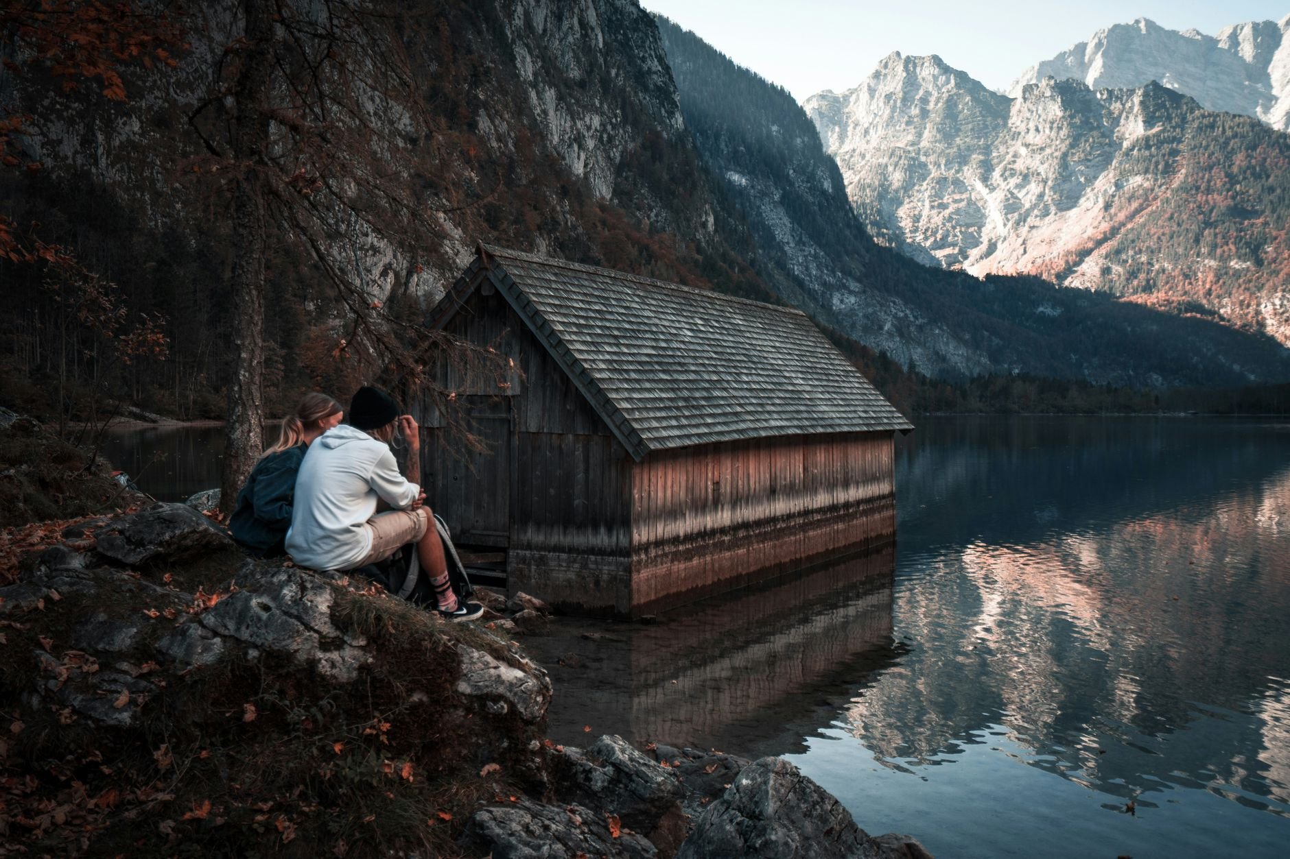 Serene scene of a couple sitting by a lake with rocky mountain views and a wooden cabin reflection.